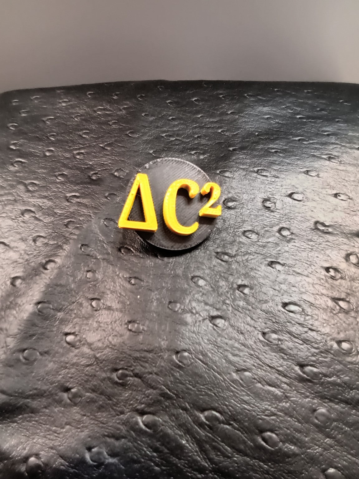 Black round Delta C squared badge with raised gold lettering